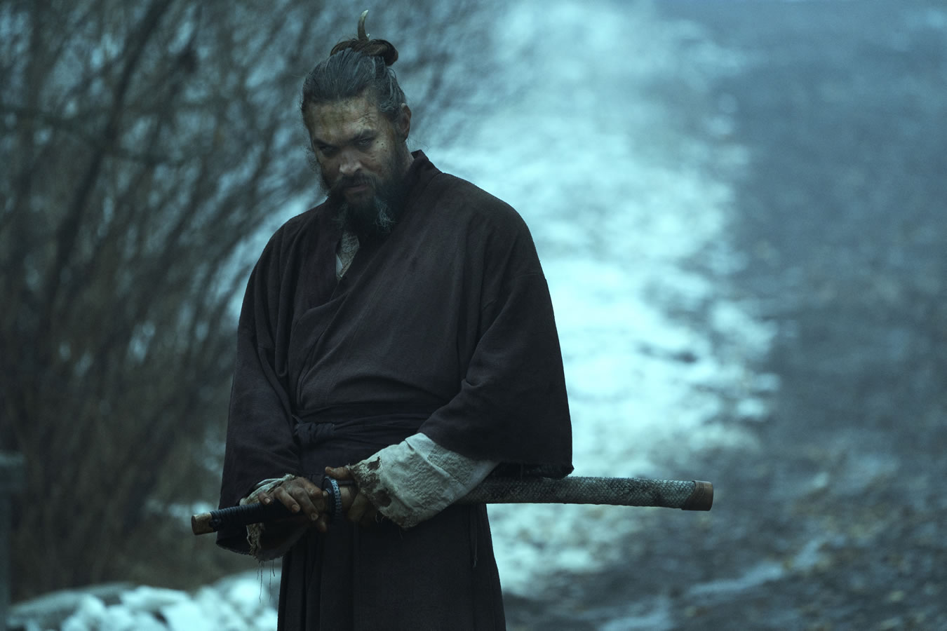 Jason Momoa, hair is in a top knot, wearing a gi holding a sword in the woods. Image courtesy of Apple